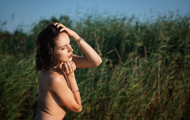 Concepto de naturaleza, belleza, juventud y estilo de vida saludable. Hermosa joven mujer desnuda sobre fondo de naturaleza. Joven mujer desnuda en la orilla del mar entre altas cañas verdes