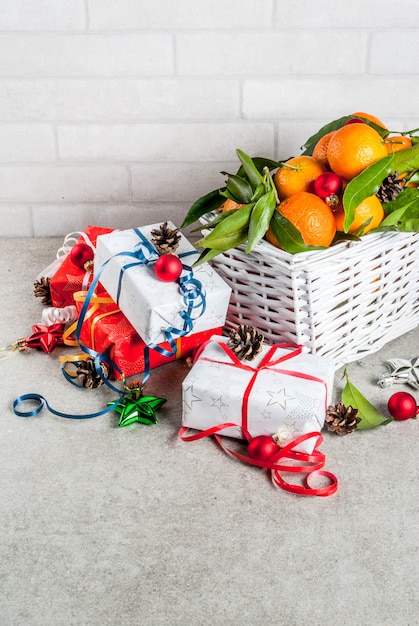 Concepto de Navidad y Año Nuevo Mandarinas frescas con hojas verdes en una cesta blanca Decoración navideña y cajas de regalo en mesa gris