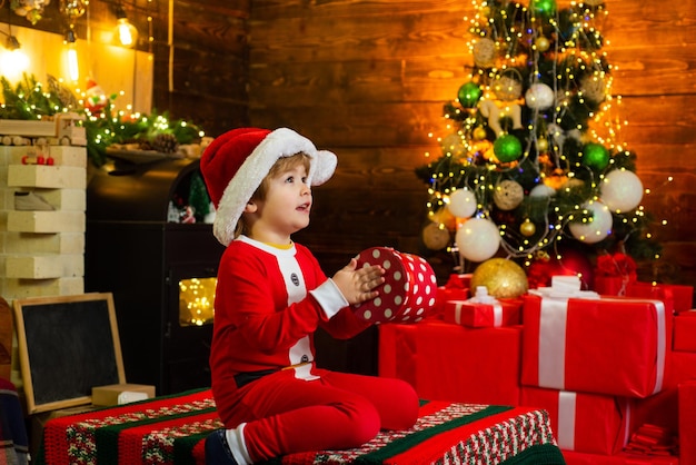 Concepto de Navidad de año nuevo. Niño feliz vestido con ropa de invierno piensa en Santa cerca del árbol de Navidad. Niño lindo alegre abriendo un regalo de Navidad.