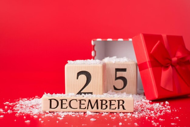 Concepto de navidad. Cerrar foto del calendario de cubo de madera que muestra la fecha de Navidad y una caja de regalo roja abierta con cinta roja aislada sobre fondo rojo con nieve