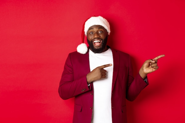 Concepto de Navidad, fiesta y vacaciones. Alegre hombre negro sonriendo, señalando con el dedo a la derecha y mostrando publicidad, de pie sobre fondo rojo.