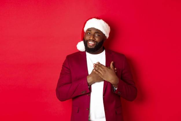 Concepto de Navidad, fiesta y vacaciones. Hombre afroamericano agradecido con sombrero de santa diciendo gracias, tomados de la mano en el corazón y sonriendo, sintiéndose conmovido, de pie contra el fondo rojo.