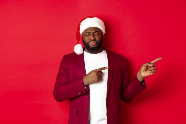 Concepto de Navidad, fiesta y vacaciones. Hombre afroamericano molesto que mira sombrío, cierra los ojos y suspira, señala con el dedo a la derecha, de pie en el sombrero de santa contra el fondo rojo.