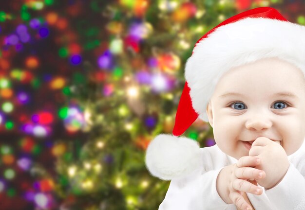 concepto de navidad, infancia, infancia y personas - bebé feliz con sombrero de santa durante las vacaciones luces de fondo