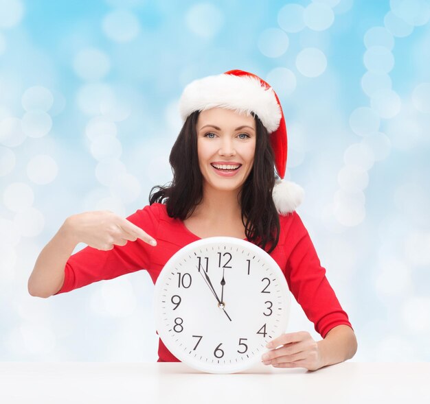 concepto de navidad, invierno, vacaciones, tiempo y personas - mujer sonriente con sombrero de ayudante de santa y vestido rojo con reloj sobre fondo de luces azules