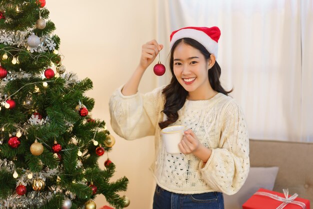 Concepto de navidad mujer asiática bebiendo una taza de café y sosteniendo una bola roja decorar en el árbol de navidad