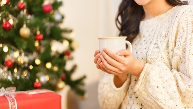 Concepto de navidad Mujer asiática joven sentada en una silla cerca del árbol de navidad y sosteniendo una taza de café