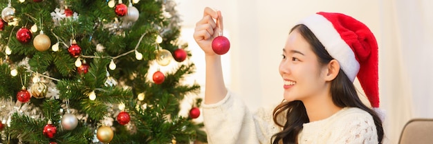 Concepto de navidad Mujer asiática joven en sombrero de santa sosteniendo bola roja para decorar en árbol de navidad