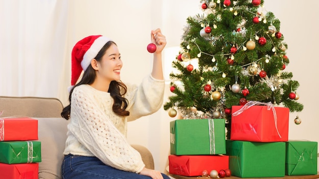 Concepto de navidad Mujer asiática joven en sombrero de santa sosteniendo bola roja para decorar en árbol de navidad