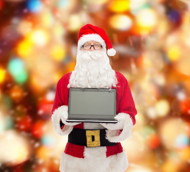 concepto de navidad, publicidad, tecnología y personas - hombre disfrazado de santa claus con computadora portátil sobre fondo de luces rojas