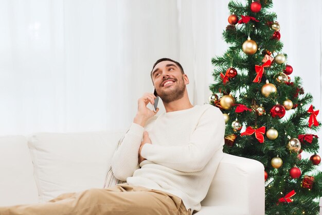 concepto de navidad, tecnología, personas y vacaciones - hombre sonriente llamando al teléfono inteligente en casa