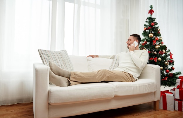 concepto de navidad, tecnología, personas y vacaciones - hombre sonriente llamando al teléfono inteligente en casa