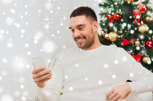 concepto de navidad, tecnología, personas y vacaciones - hombre sonriente con mensaje de texto de teléfono inteligente en casa