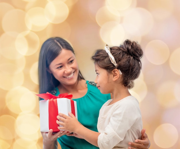 Foto concepto de navidad, vacaciones, celebración, familia y personas - madre feliz y niña con caja de regalo sobre fondo de luces beige