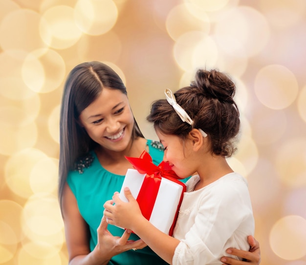 concepto de navidad, vacaciones, celebración, familia y personas - madre feliz y niña pequeña con caja de regalo sobre fondo de luces beige