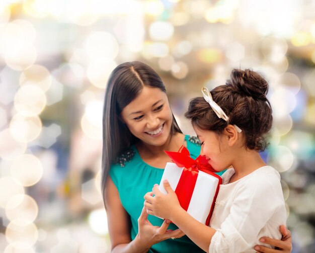 concepto de navidad, vacaciones, celebración, familia y personas - madre feliz y niña pequeña con caja de regalo sobre fondo de luces