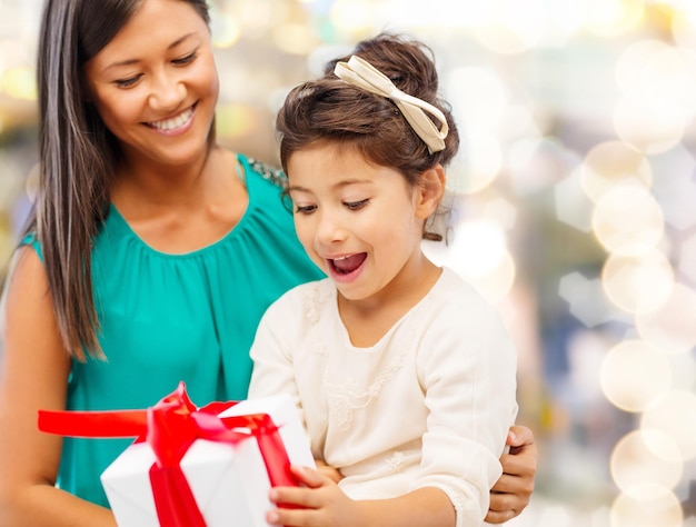 concepto de navidad, vacaciones, celebración, familia y personas - madre feliz y niña pequeña con caja de regalo sobre fondo de luces