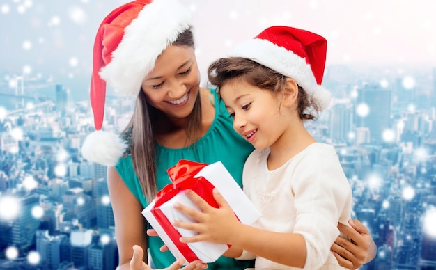 concepto de navidad, vacaciones, celebración, familia y personas - madre feliz y niña pequeña con sombreros de ayudante de santa con caja de regalo sobre el fondo de la ciudad nevada