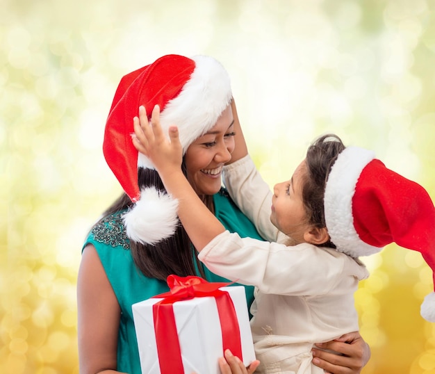 concepto de navidad, vacaciones, celebración, familia y personas - madre feliz y niña con sombreros de ayudante de santa con caja de regalo sobre fondo de luces amarillas