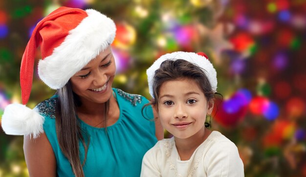 concepto de navidad, vacaciones, familia, infancia y personas - madre feliz y niña pequeña con sombreros de santa sobre fondo de luces