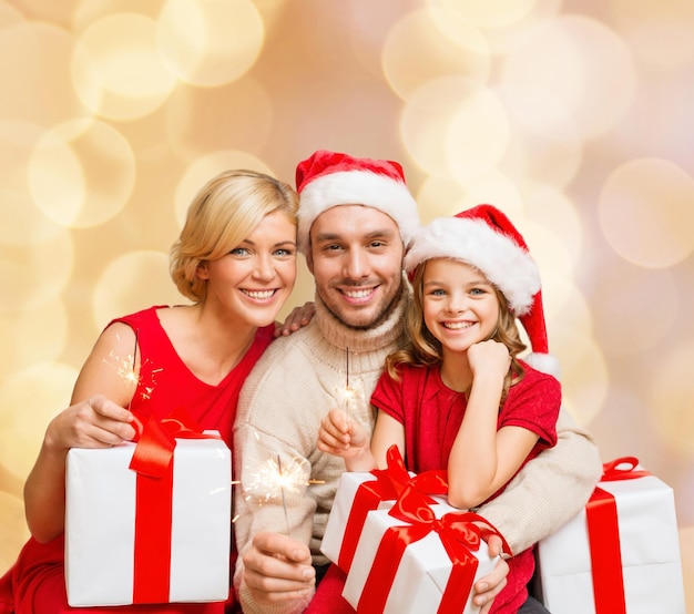 concepto de navidad, vacaciones, familia y personas - feliz madre, padre y niña en sombreros de ayudante de santa con cajas de regalo sobre fondo de luces beige