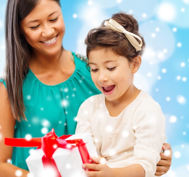 concepto de navidad, vacaciones, familia y personas - madre feliz y niña con caja de regalo sobre fondo de luces azules