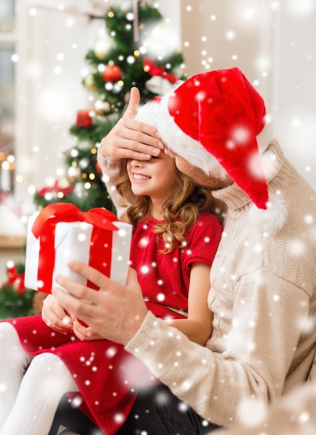 concepto de navidad, vacaciones, familia y personas - padre e hija sonrientes con sombreros de santa sosteniendo una caja de regalo y cubriendo los ojos en casa