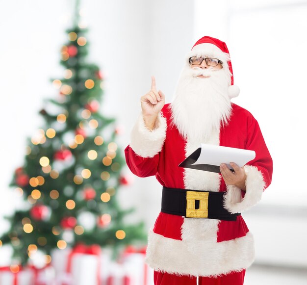 concepto de navidad, vacaciones, gestos y personas - hombre disfrazado de santa claus con bloc de notas apuntando con el dedo hacia arriba sobre la sala de estar con fondo de árbol