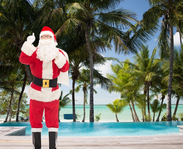 concepto de navidad, vacaciones, gestos, viajes y personas - hombre disfrazado de santa claus con una bolsa apuntando con el dedo sobre la playa tropical y el fondo de la piscina