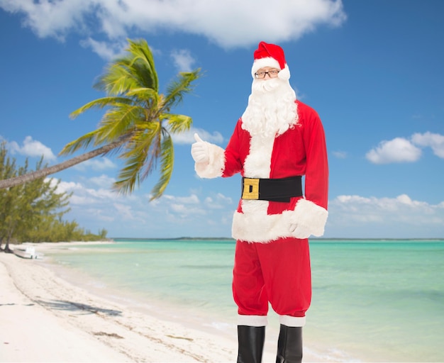 concepto de navidad, vacaciones, gestos, viajes y personas- hombre disfrazado de santa claus mostrando los pulgares hacia arriba sobre el fondo de la playa tropical