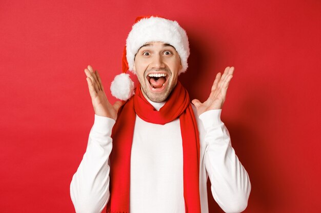 Concepto de Navidad, vacaciones de invierno y celebración. Imagen de un hombre guapo mirando sorprendido por la oferta de promoción de año nuevo, sonriendo asombrado, de pie sobre fondo rojo.