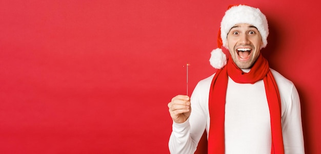 Concepto de navidad, vacaciones de invierno y celebración. Primer plano de un hombre feliz y guapo con sombrero y bufanda de santa, divirtiéndose en la fiesta de año nuevo, sosteniendo un bengala y sonriendo, fondo rojo.