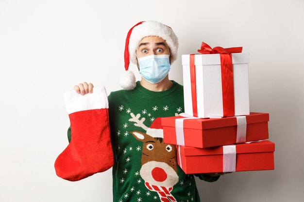 Concepto de Navidad y vacaciones de invierno. Hombre feliz en mascarilla y gorro de Papá Noel trayendo regalos, sosteniendo un calcetín de Navidad y cajas de regalo, de pie sobre fondo blanco.