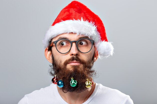 Concepto de Navidad, vacaciones, peluquería y estilo - joven apuesto hombre barbudo de santa claus con muchos pequeños adornos navideños en barba larga.