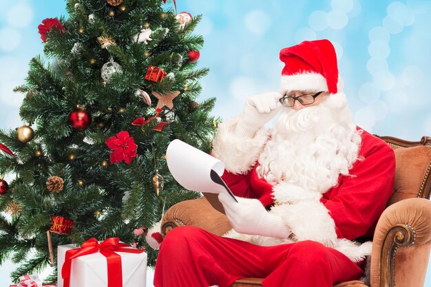 concepto de navidad, vacaciones y personas - hombre disfrazado de santa claus con bloc de notas y árbol de navidad sentado en un sillón sobre fondo de luces azules
