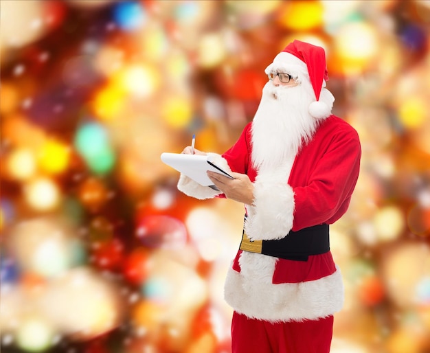 concepto de navidad, vacaciones y personas - hombre disfrazado de santa claus con bloc de notas y bolígrafo sobre fondo de luces rojas