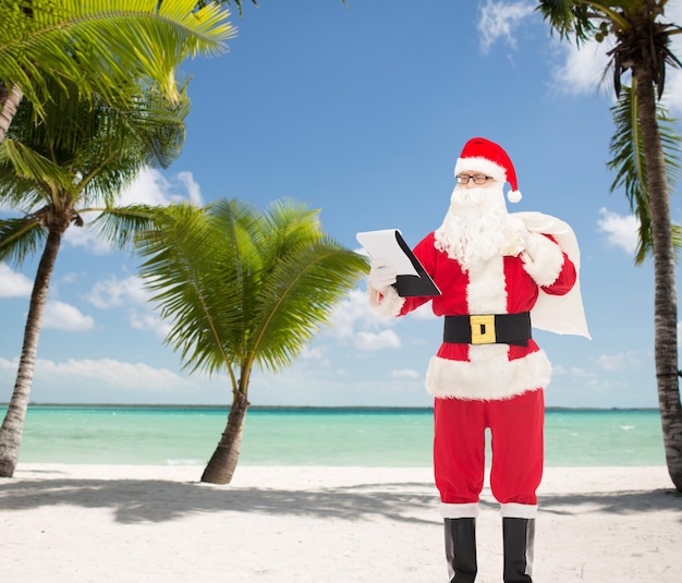 concepto de navidad, vacaciones y personas - hombre disfrazado de santa claus con bloc de notas y bolsa sobre fondo de playa tropical