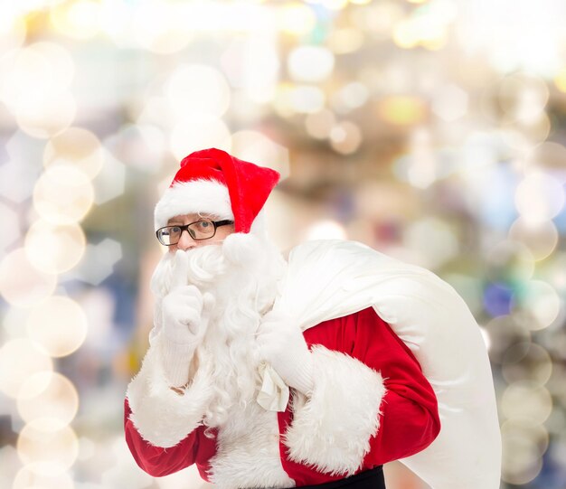 concepto de navidad, vacaciones y personas - hombre disfrazado de santa claus con bolsa haciendo gesto de silencio sobre fondo de luces