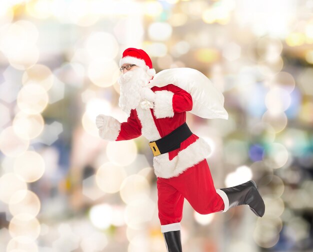 concepto de navidad, vacaciones y personas - hombre disfrazado de santa claus corriendo con bolsa sobre fondo de luces