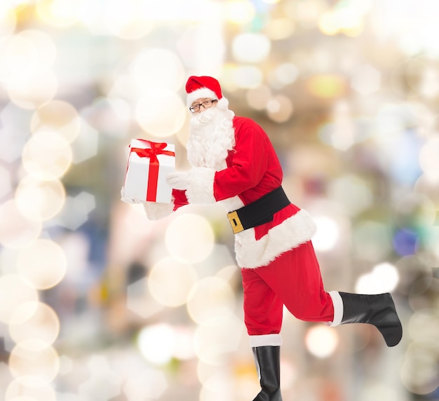 concepto de navidad, vacaciones y personas - hombre disfrazado de santa claus corriendo con caja de regalo sobre fondo de luces