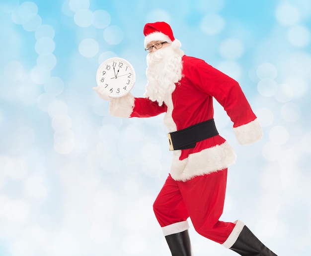 concepto de navidad, vacaciones y personas - hombre disfrazado de santa claus corriendo con un reloj que muestra doce sobre fondo de luces azules