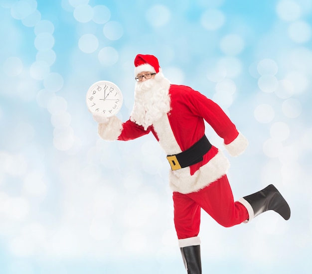 concepto de navidad, vacaciones y personas - hombre disfrazado de santa claus corriendo con un reloj que muestra doce sobre fondo de luces azules