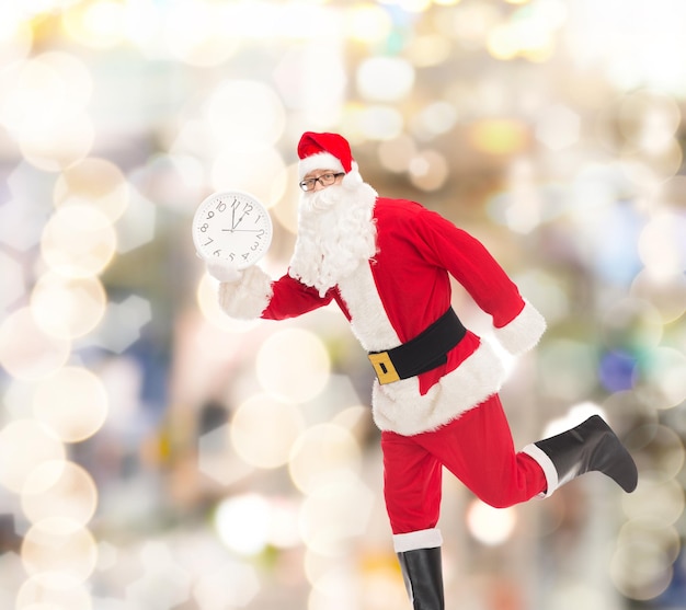 concepto de navidad, vacaciones y personas - hombre disfrazado de santa claus corriendo con un reloj que muestra doce sobre fondo de luces