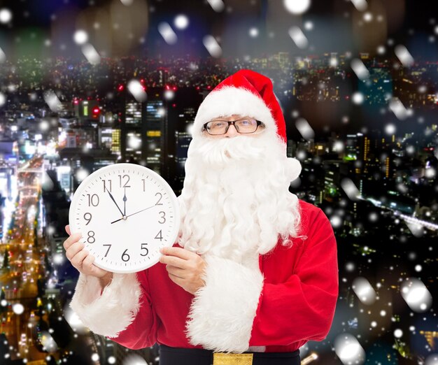 concepto de navidad, vacaciones y personas - hombre disfrazado de santa claus con reloj que muestra doce sobre fondo de ciudad de noche nevada