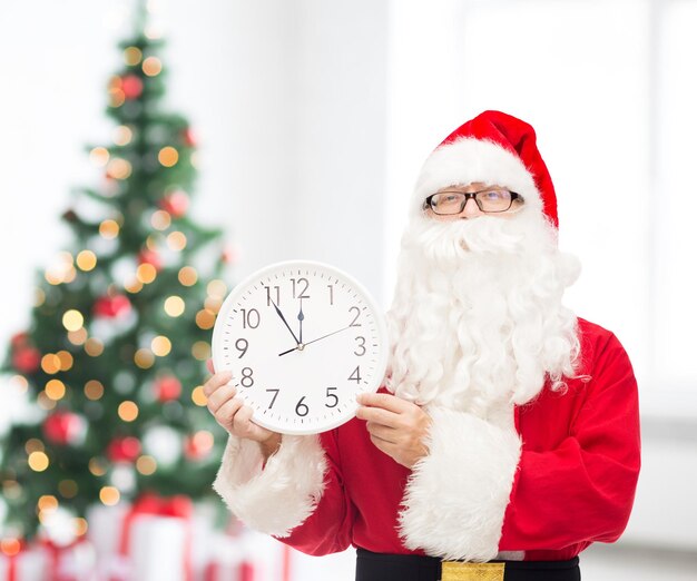 concepto de navidad, vacaciones y personas - hombre disfrazado de santa claus con reloj que muestra doce sobre la sala de estar y el fondo del árbol