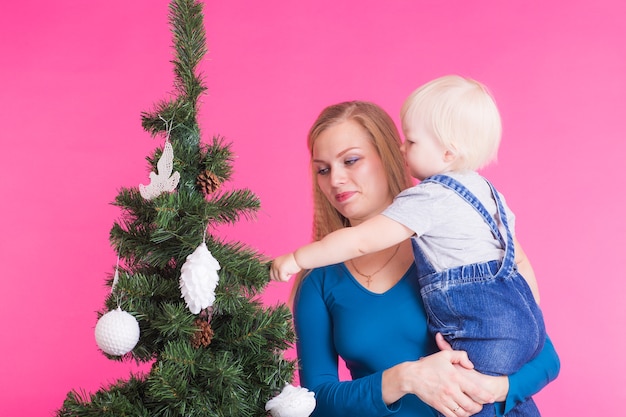 Concepto de Navidad, vacaciones y personas - mujer y su hijo cerca del árbol de Navidad sobre fondo rosa
