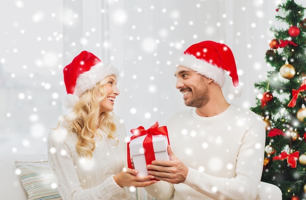 concepto de navidad, vacaciones y personas - pareja feliz con sombreros de santa con caja de regalo sentada en el sofá en casa