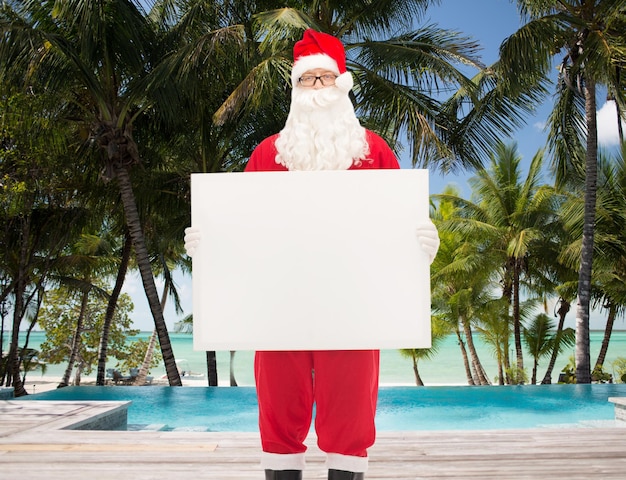 concepto de navidad, vacaciones, publicidad, viajes y personas - hombre disfrazado de santa claus sosteniendo una cartelera blanca en blanco sobre la piscina en el fondo de la playa tropical