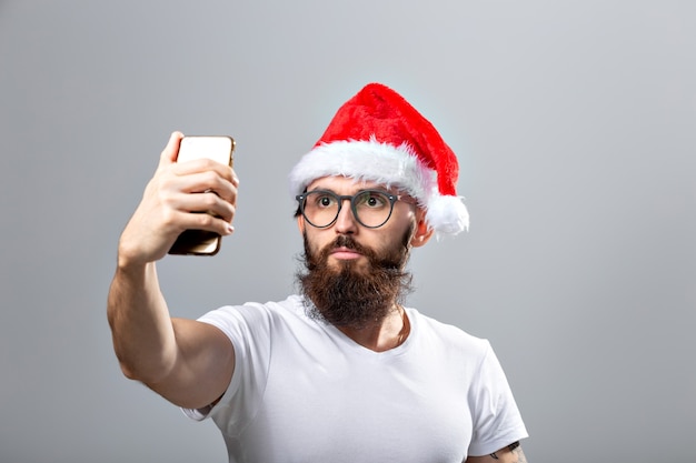 Concepto de Navidad, vacaciones, tecnología y personas - hombre barbudo guapo con sombrero de santa tomando selfie