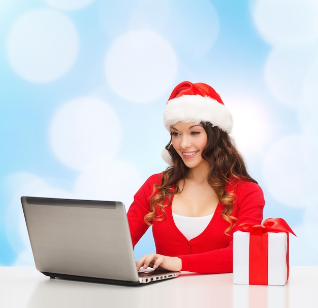 concepto de navidad, vacaciones, tecnología y personas: mujer sonriente con sombrero de ayudante de santa con caja de regalo y computadora portátil sobre fondo de luces azules